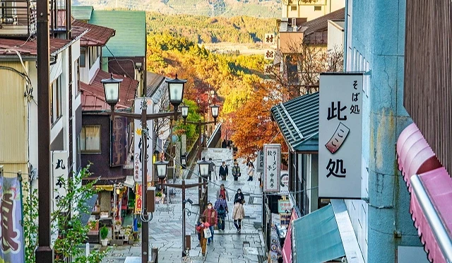從高處俯瞰伊香保石段街,石階向下延伸,兩旁為店舖與建築,行人散步其間,背景為秋色山景